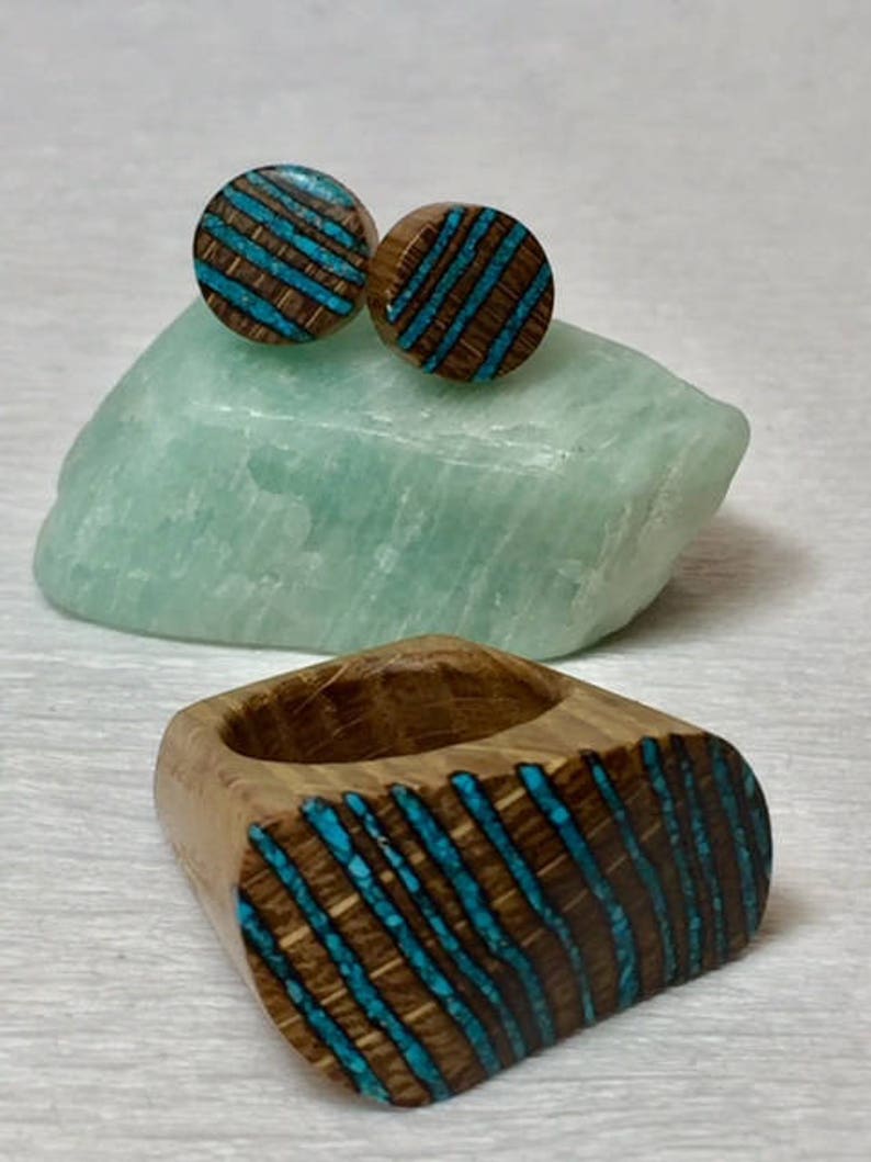 Puede incluir: Un conjunto de pendientes de madera y un anillo con incrustaciones de turquesa. Los pendientes son redondos y el anillo tiene forma ovalada. Las incrustaciones de turquesa tienen un patr&oacute;n de rayas.