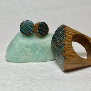 Puede incluir: Un anillo de madera y un par de pendientes con incrustaciones de turquesa. El anillo es una banda ancha con forma cuadrada y los pendientes son redondos. La madera es de color marr&oacute;n claro y la incrustaci&oacute;n de turquesa es de un azul brillante.