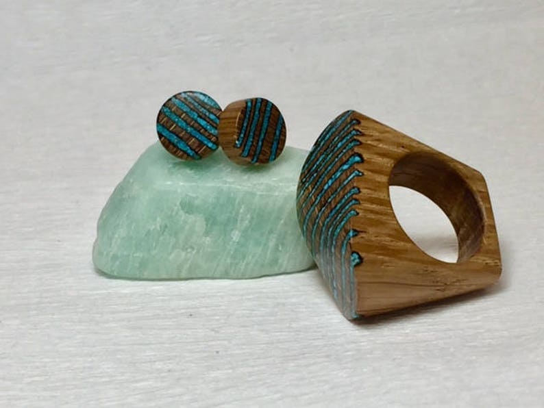 Puede incluir: Un anillo de madera y un par de pendientes con incrustaciones de turquesa. El anillo es una banda ancha con forma cuadrada y los pendientes son redondos. La madera es de color marr&oacute;n claro y la incrustaci&oacute;n de turquesa es de un azul brillante.
