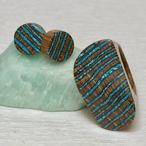 Puede incluir: Un conjunto de pendientes de madera y un anillo con incrustaciones de turquesa. Los pendientes son redondos y el anillo tiene forma ovalada. La incrustaci&oacute;n de turquesa tiene un patr&oacute;n de rayas.