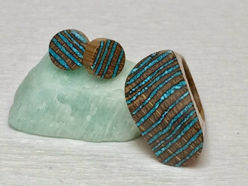 Puede incluir: Un conjunto de pendientes de madera y un anillo con incrustaciones de turquesa. Los pendientes son redondos y el anillo tiene forma ovalada. La incrustaci&oacute;n de turquesa tiene un patr&oacute;n de rayas.