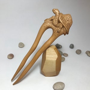 May include: A wooden hair stick with a carved turtle design. The turtle is holding a snake in its mouth. The stick is resting on a wooden block.
