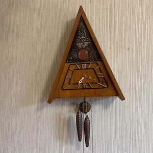 Könnte beinhalten: Dreieckige Kuckucksuhr aus Holz mit dekorativem Messingaufsatz und Zifferblatt mit römischen Ziffern. Die Uhr hat zwei tannenzapfenförmige Gewichte und eine Metallkette. Die Uhr ist an einer hellen Wand befestigt.