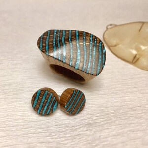 Puede incluir: Un conjunto de joyas de madera que incluye un anillo y pendientes. El anillo y los pendientes est&aacute;n hechos de madera oscura con rayas azul turquesa.
