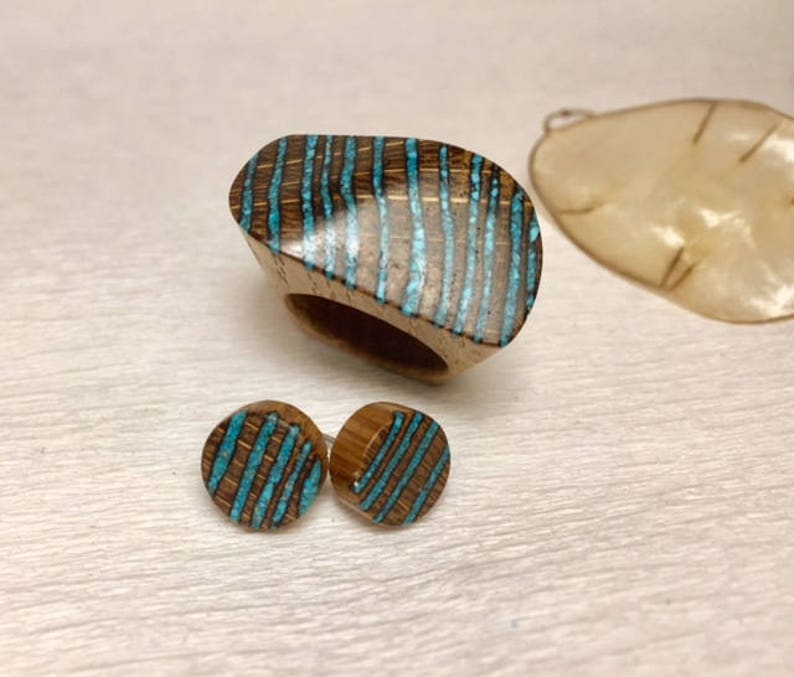 Puede incluir: Un conjunto de joyas de madera que incluye un anillo y pendientes. El anillo y los pendientes est&aacute;n hechos de madera oscura con rayas azul turquesa.