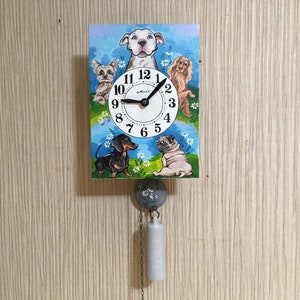 May include: A wooden clock with a blue background and a white clock face. The clock face has black Roman numerals and a black hour and minute hand. The clock is surrounded by five cartoon dogs, including a dachshund, a pug, a Yorkshire terrier, and two other dogs. The clock is hanging from a chain with a white weight.