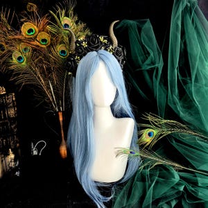 May include: A black headband with horns and black roses. The headband is worn on a mannequin with long, light blue hair. The mannequin is posed against a green fabric background.
