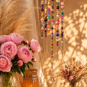May include: A hanging mobile featuring a pink, textured top disc with multiple strands of multicolored beads in various sizes and finishes, including metallic, translucent, and opaque spheres, ending with a clear faceted crystal prism.