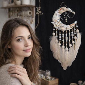 May include: A crescent moon-shaped dreamcatcher made of seashells, beads, and feathers. The dreamcatcher is cream-colored with gold accents and hangs against a black background. The feathers are long and flowing.