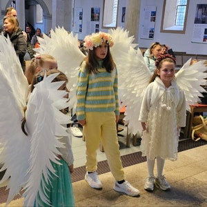 Op de afbeelding: Drie kinderen verkleed als engelen met witte vleugels en witte jurken. De kinderen staan in een kerk met een houten vloer en banken op de achtergrond.