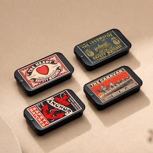 Può includere: Quattro scatole di fiammiferi rettangolari nere con etichette in stile vintage. Le etichette presentano disegni come un cuore, una locomotiva, un'ancora e una carovana. Il testo sulle scatole recita "Safety Matches" e altre frasi.