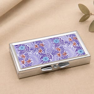 May include: A rectangular silver pill box with a floral design. The lid features a pattern of purple leaves, blue flowers, and orange accents. The box is closed and sits on a beige surface.