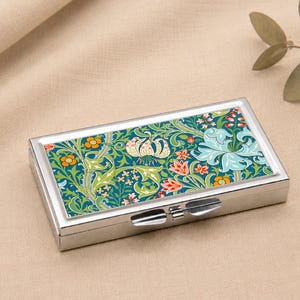 May include: A rectangular silver pill box with a colorful floral design. The box features a hinged lid and a vibrant pattern of green leaves, orange flowers, and blue and white lilies. The background is a neutral beige.