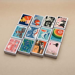 May include: A selection of vintage matchboxes with various illustrations. Each box displays a different design, such as a portrait, an octopus, and a mountain landscape. The matchboxes are arranged on a neutral background.