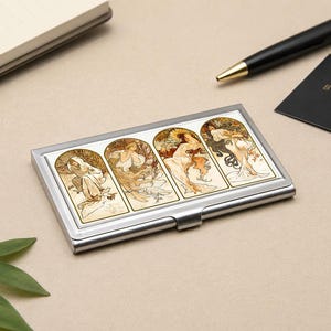 May include: A silver business card holder with four Art Nouveau illustrations of women in flowing gowns. The artwork has cream, gold, and brown tones. A pen and notepad are in the background. The holder is made of metal.