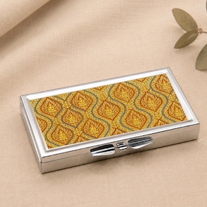 May include: A rectangular silver metal case featuring a top panel with a repeating gold and dark green intricate pattern. The case has a hinged lid with a central metal latch mechanism on the front edge, designed for holding small items securely.