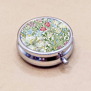 May include: A round, silver pill box with a floral design on the lid. The lid features a colorful pattern of flowers and leaves in shades of green, pink, blue, and white. The pill box has a small, silver button on the side.