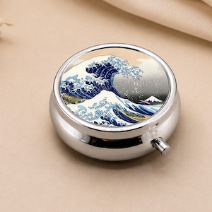 May include: A circular, silver pill box with a print of Hokusai's "The Great Wave off Kanagawa." The artwork shows a large ocean wave and a mountain. The pill box has a small, silver button for opening.