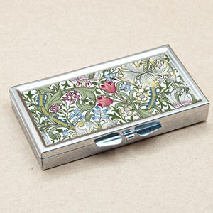 May include: A silver pill box with a floral design featuring lilies, leaves, and flowers in shades of green, yellow, white, pink, and blue.