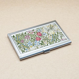 May include: A rectangular business card holder with a silver metal frame. The top surface has a colourful floral pattern with red, pink, green, blue, and white elements. The design is reminiscent of vintage botanical illustrations.