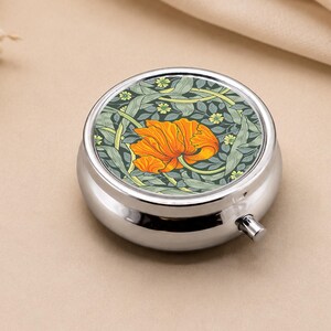 Peut inclure: Une boîte à pilules ronde et argentée avec un motif floral sur le couvercle. Le motif présente une fleur orange avec des feuilles vertes et des accents jaunes. La boîte à pilules est sur un fond neutre.