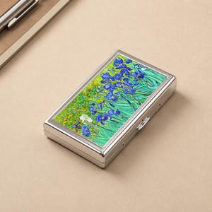 May include: A rectangular, silver metal case with a hinged lid. The top features a vibrant painting of blue irises in a field of green and yellow, reminiscent of Van Gogh's style. The case is likely for holding small items.