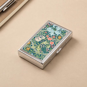 Könnte beinhalten: Ein rechteckiges, silberfarbenes Etui mit floralem Design. Das Etui weist ein detailliertes Muster aus grünen, blauen, orangefarbenen und cremefarbenen Blumen und Blättern auf dunklem Teal-Hintergrund auf. Das Etui ist geschlossen und hat einen kleinen Verschluss.