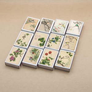 Könnte beinhalten: Zwölf rechteckige Schachteln mit Aquarellmalereien von Vögeln und Blumen. Jede Schachtel hat einen weißen Rahmen und einen beigen Hintergrund. Die Kunstwerke zeigen Vögel, Blüten und Blätter in Rosa-, Grün- und Blautönen.