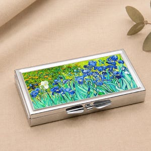 Può includere: Un portapillole rettangolare argentato con coperchio incernierato. La parte superiore presenta un dipinto colorato di iris blu e fogliame verde. La scatola è appoggiata su una superficie beige, con un rametto di foglie sullo sfondo.