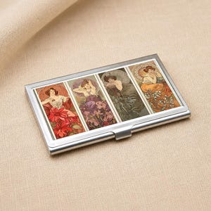 May include: A rectangular silver metal business card case featuring four vertical panels with illustrations of figures surrounded by floral motifs in shades of red, purple, green, and orange, with a polished metallic frame and a hinged closure.
