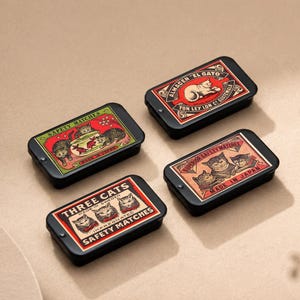 Può includere: Quattro scatole di fiammiferi rettangolari nere con etichette vintage a tema gatto. Le etichette presentano illustrazioni di gatti e testo come "THREE CATS SAFETY MATCHES" e "ALMACEN EL GATO". Le scatole sono disposte su una superficie dai toni neutri.