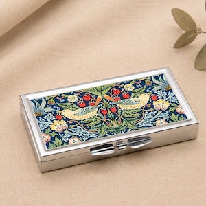 May include: A rectangular, silver pill box with a decorative lid. The lid features a detailed floral and bird design in shades of blue, green, yellow, and red. The box is closed with a silver clasp.
