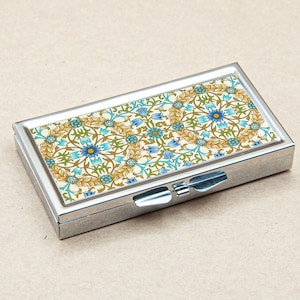 May include: A rectangular, silver-toned pill box with a floral design on the lid. The pattern features blue, green, and gold flowers and leaves against a cream background. The box has a hinged closure.