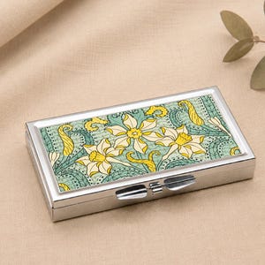May include: A rectangular, silver pill box with a floral design on the lid. The design features yellow and white flowers with green and blue accents. The box is closed with a silver clasp.