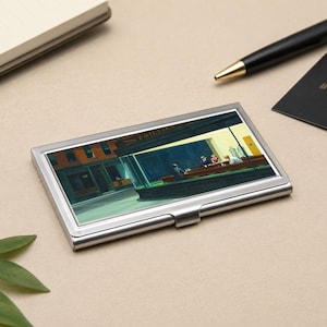 May include: A silver business card holder featuring a print of Edward Hopper's "Nighthawks" painting. The artwork depicts a diner scene with figures inside. A black pen, a notepad, and a business card are also in the image.
