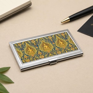 May include: A rectangular metal business card holder featuring a repeating pattern of golden Buddha figures seated within ornate pointed arches against a deep blue background with intricate gold floral filigree details and a polished silver frame.
