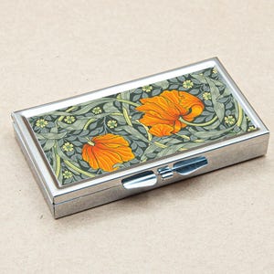 May include: A rectangular, silver-toned pill box with a hinged lid. The top features a floral design with orange flowers and green foliage on a dark green background. The box is closed.