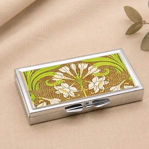 May include: A rectangular silver pill box with a floral design. The lid has a pattern of white flowers, green leaves, and a brown background. The box is closed with a silver clasp and rests on a tan surface.
