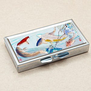 Puede incluir: Un pastillero rectangular plateado con tapa abisagrada. La parte superior presenta una colorida ilustración de peces koi nadando en un estanque con agua azul y detalles florales. Los peces son rojos, blancos y naranjas.