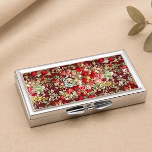 May include: A rectangular, silver pill box with a floral design on the lid. The pattern shows red, pink, and yellow flowers on a dark red backdrop. The box is secured with a silver clasp.