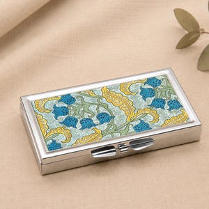 May include: A rectangular, silver pill box with a hinged lid. The top features a floral design in blue, yellow, and green. The box is on a beige surface.