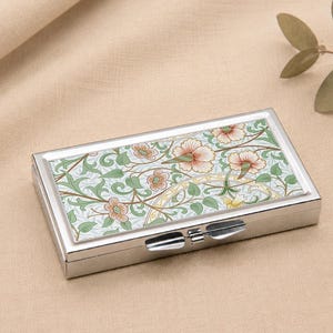 May include: A rectangular silver pill box with a floral pattern on the lid. The design includes peach-coloured flowers, green leaves, and a light blue backdrop. The box is secured with a silver clasp.