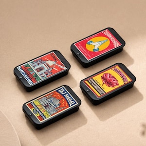 Può includere: Quattro scatole di fiammiferi rettangolari nere con etichette colorate in stile vintage. Le etichette presentano illustrazioni di un Taj Mahal, un fiore di loto, un razzo e un Taj Mahal. Le scatole di fiammiferi sono disposte su una superficie neutra.