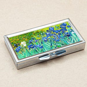 May include: A silver pill box with a hinged lid featuring a colourful painting of blue irises on a green background.