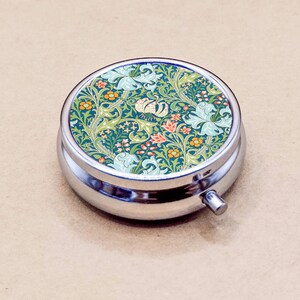 May include: A circular, silver pill box with a decorative floral pattern on the lid. The lid displays a design of green, blue, orange, and white flowers and leaves. The pill box has a small silver button for opening.