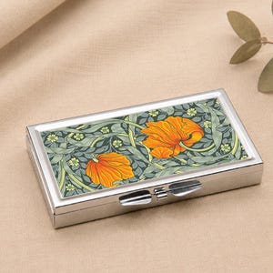 Op de afbeelding: Een rechthoekige zilveren pillendoos met een bloemenontwerp. De deksel heeft een patroon van oranje bloemen en groene bladeren op een donkergroene achtergrond. De doos is gesloten met een zilveren sluiting.