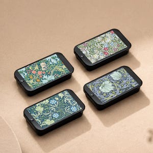 May include: Four small, black rectangular boxes with floral patterned lids. The lids feature various designs with green, blue, red, and white flowers and foliage. The boxes are arranged on a light brown surface.