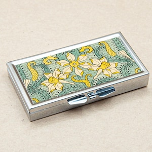 May include: A silver pill box with a floral design. The design features yellow flowers with green leaves on a light blue background.