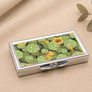Op de afbeelding: Een rechthoekige zilveren pillendoos met een scharnierend deksel. De bovenkant heeft een bloemenontwerp van groene waterleliebladeren en gele bloemen op een donkere achtergrond. De doos staat op een neutrale ondergrond.