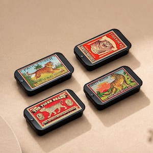 May include: Four vintage-style matchboxes with black metal cases. Each box features a different tiger illustration and text, including "PALM & TIGER", "TIGERHEAD", "THE TIGER BRAND", and "SAFETY MATCH". The boxes are arranged on a neutral surface.